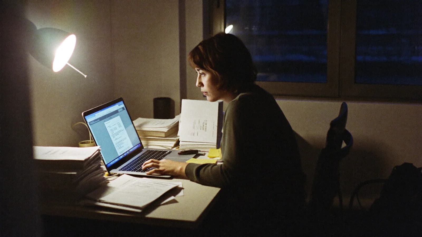 A small business owner working alone at a desk late at night, laptop open beside stacks of paperwork and invoices.